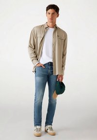 Casual outfit med en beige langærmet skjorte, hvid t-shirt, blå jeans og grøn kasket, sammen med hvide og beige sneakers.
