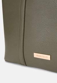 Olīvu zaļš rokassomu, izgatavota no teksturēta materiāla, ar zelta zīmola plāksni un akcentētiem šuvēm, taisnstūra forma.