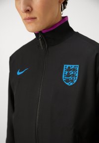 Black Nike jacket with a high collar, featuring a blue England badge on the chest and a blue logo on the left shoulder. Smooth texture.