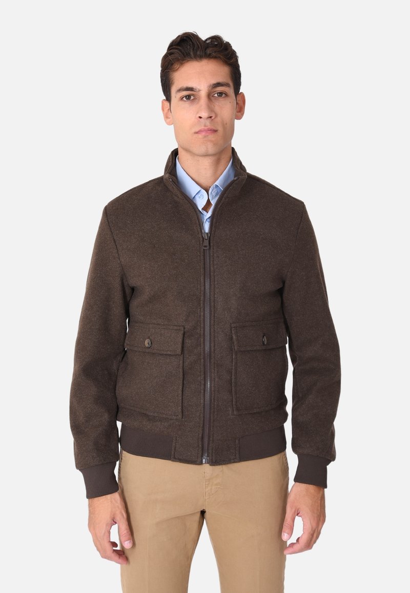 Giacca bomber marrone con chiusura a zip, due tasche frontali, polsini e orlo a coste, realizzata in un tessuto testurizzato. Indossata sopra una camicia azzurro chiaro.