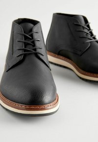 Botas chukka negras de cuero con un acabado texturizado, diseño de cordones, entresuela crema y costuras de acento marrón a lo largo del borde de la suela.