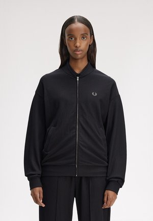 Mujer de cabello lacio con chaqueta negra con cremallera y pantalones a juego, de pie frente a un fondo claro liso, mirando hacia adelante.