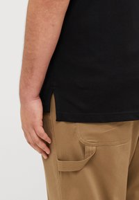 Μαύρο κοντομάνικο πουκάμισο με καμπυλωτό στρίφωμα, το οποίο συνδυάζεται με μπεζ παντελόνι που διαθέτει πλαϊκή τσέπη και ορατές ραφές. Χέρι resting πάνω στο παντελόνι.