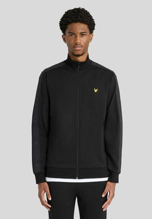 Young man with short curly hair wearing a black zip-up jacket with a small yellow bird logo on the left chest and black pants.