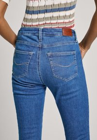 Jeans de mezclilla azul con cinco bolsillos, una textura suave y un parche de cuero marrón en la cinturilla. Diseño clásico de corte ajustado.