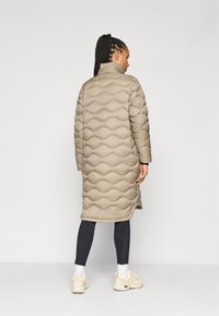 Beige gewatteerde lange jas met een hoge kraag, met een getextureerd golvend ontwerp. G gedragen over donkere leggings en beige sneakers.