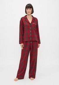 Czerwony zestaw piżamowy w kratkę, wykonany z miękkiego materiału, z górą zapinaną na guziki i kieszonką na piersi oraz luźnymi spodniami, obydwa elementy z kontrastującymi czarnymi liniami.