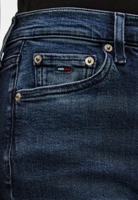 Tuvplāns tumši zilām džinsa biksēm ar metāla kniežiem un mazu sarkanu, baltu un zilu zīmolu uz monētu kabatas.