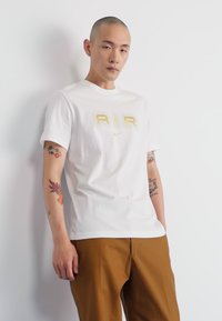 Bijela pamučna majica s zlatnim logotipom "AIR" i Nikeovim swooshom. Uparena s bež hlačama, prikazana na svijetloj pozadini.