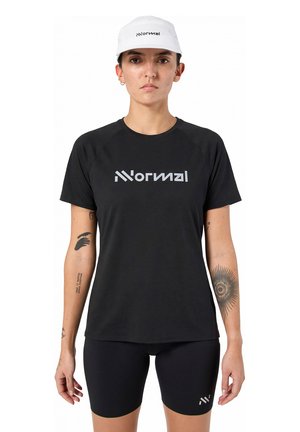 Vrouw draagt een witte pet en een zwart sportoutfit met het logo "Normal", staat met zichtbare tatoeages op beide armen tegen een effen achtergrond.