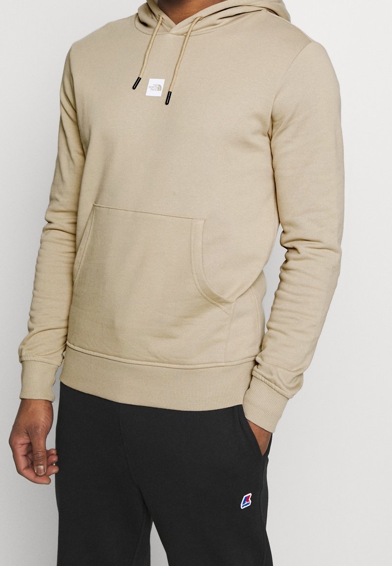 Personne portant un sweat à capuche beige avec poche avant et un pantalon de jogging noir avec un logo sur la cuisse, debout devant un fond clair uni.