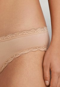 Beige ondergoed met kantafwerking en een gladde, zachte textuur. Golvende randen langs de tailleband voegen een delicate detail toe.