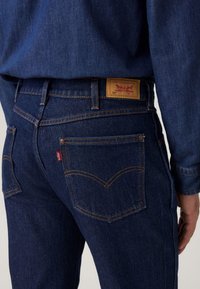 Jeans de mezclilla azul oscuro con costuras naranjas, que presentan la etiqueta con el logo de Levi's en la cintura trasera y un diseño en el bolsillo trasero.