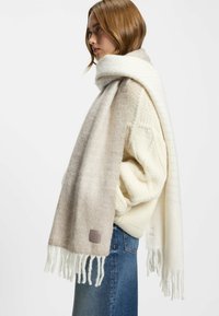 Beige og hvidt tørklæde lavet af blød stof, med en gradient design, frynsede kanter og et lille brandmærke i den ene ende.