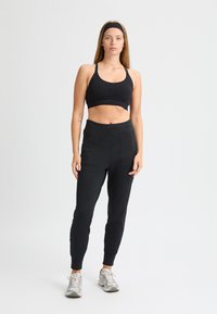 Sujetador deportivo negro con detalle fruncido, combinado con pantalones jogger negros ajustados que cuentan con bolsillos laterales. El modelo lleva zapatillas deportivas gris claro.