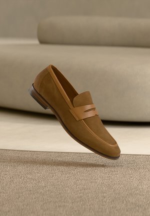 Brown suede men's loafer with leather strap detail, displayed above a beige carpet and in front of a light beige sofa.