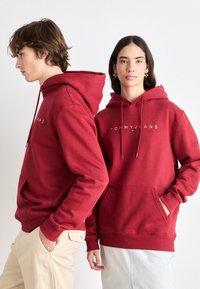 Sudadera roja hecha de una mezcla de algodón, con un bolsillo delantero y capucha con cordón. En la parte frontal aparece "TOMMY JEANS" en letras doradas.