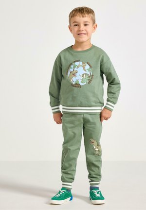 Completo di felpa e pantaloni tuta verde realizzato in tessuto morbido. Presenta grafiche di dinosauri e polsini a righe. Abbinato a sneaker verdi.