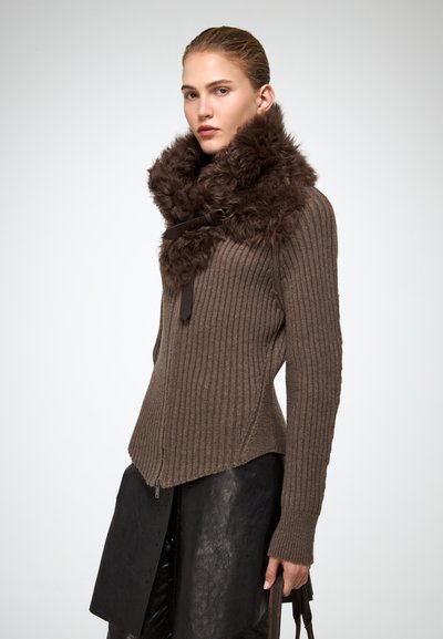 Donna che indossa un maglione a coste marrone con un ampio colletto in pelliccia marrone scuro e una gonna in pelle nera, mentre tiene in mano manici marroni su uno sfondo neutro.
