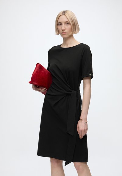 Femme aux cheveux blonds courts portant une robe noire avec un nœud sur le côté, tenant une pochette rouge texturée sur un fond clair uni.