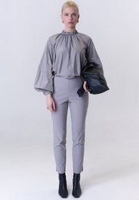 Hellgraue Bluse mit hohem, gerafftem Kragen und voluminösen Ärmeln, kombiniert mit engen hellgrauen Hosen und schwarzen Stiefeletten mit Absatz.