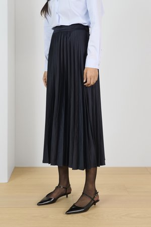 Faltenrock in Marineblau aus leichtem Material, kombiniert mit schwarzen spitz zulaufenden Ballerinas und transparenten schwarzen Strumpfhosen, von der Taille abwärts auf einem Holzboden gezeigt.