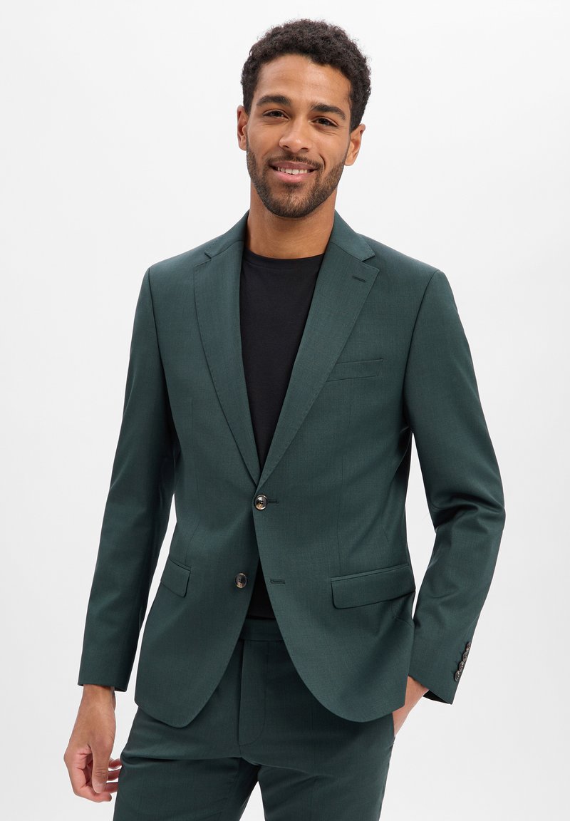 Dunkelgrüner, maßgeschneiderter Blazer mit fallendem Revers, Ein-Knopf-Verschluss und Pattentaschen, kombiniert mit einem schwarzen T-Shirt darunter.