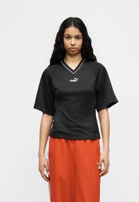 Γυναίκα όρθια, κοιτάζοντας μπροστά, φοράει μαύρο αθλητικό τοπ Puma και φαρδιά κόκκινα παντελόνια με λευκές ρίγες στα πλάγια, μπροστά σε απλό φόντο.