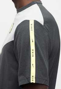 Αθλητική κοντομάνικη μπλούζα ανθρακί γκρι με λευκά πάνελ στους ώμους και κίτρινο λογότυπο Nike κατά μ ήκος του μανικιού. Κατασκευασμένη από απαλό ύφασμα.