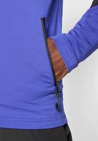 Violetsīgas garām piedurknēm blūze ar gludu apdari, kurai ir melna rāvējslēdzēja kabata sānos. Dizainā ir iekļauts melns akcents pie pleca.