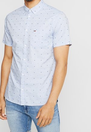 Homme portant une chemise légère à manches courtes bleu clair à motifs de petits carrés bleus et un jean bleu clair, debout devant un fond uni.