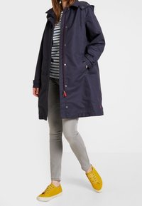 Giacca lunga blu navy con cappuccio, caratterizzata da una chiusura con zip e bottoni a pressione, abbinata a una maglietta a righe, pantaloni grigi e sneaker gialle.