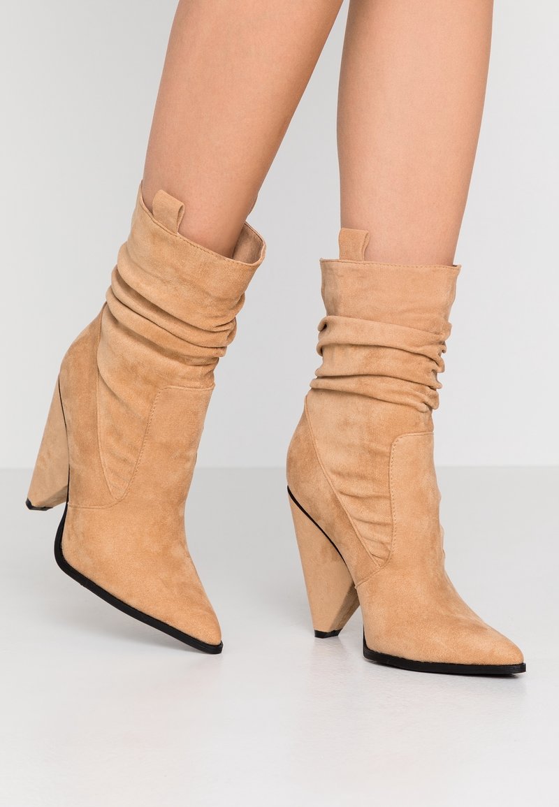 Bottes cheville en daim tan avec une tige souple, un bout pointu et un talon compensé courbé et haut. Accent de semelle extérieure noire à l'avant.