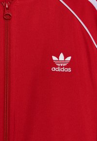 Piros, sima anyagú, cipzáros dzseki, fehér díszcsíkokkal és hímzett fehér Adidas logóval a bal mellkas területén.