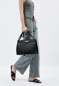 Borsa a tracolla in pelle nera con un design strutturato, dotata di chiusura con zip e hardware in tonalità oro, tenuta da una persona che indossa jeans a gamba larga di colore blu chiaro.