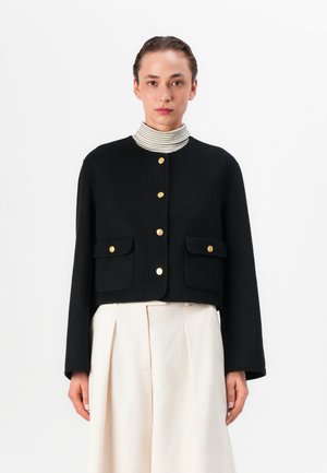 Zwarte cropped jas met gouden knoopsluiting, twee voorkruiden en een ronde halslijn, gecombineerd met een structurele witte rok.
