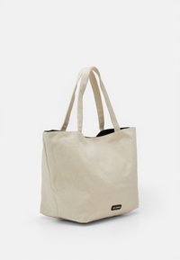 Beige kangaskassi, jossa on kaksi kantohihnaa, musta sisusta ja pieni brändätty etiketti edessä. Yksinkertainen suorakaiteen muotoinen design.