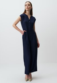 Combinaison bleu marine à manches courtes, fermeture à boutons sur le devant, tissu texturé, coupe lâche et poches latérales, accompagnée de chaussures à talons neutres.