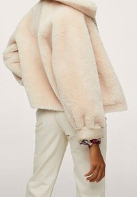 Persona vestida con una chaqueta esponjosa color crema con puños elásticos y pantalones beige claros, de pie frente a un fondo liso.