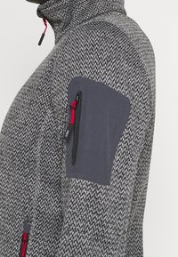 Szara kurtka w wzory, z wysokim kołnierzem, kieszenią na zamek z czerwonym akcentem oraz kontrastową szarą łatą na ramieniu dla zwiększenia trwałości.