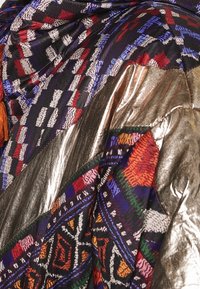 Tissu avec des motifs géométriques vibrants en rouge, bleu, blanc et orange, combinés à des sections métalliques dorées brillantes et des détails en pampilles.