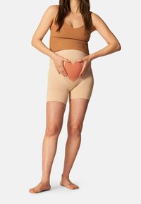 Pantaloni shorts beige a vita alta con una texture a forma di cuore ritagliata nella parte anteriore. Modella che indossa un top a canotta marrone a costine, scalza, con le mani sui fianchi.