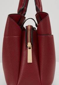 Rote Lederhandtasche mit strukturierter Oberfläche, einer goldfarbenen Reißverschlusstasche, goldenen Akzenten und zwei Griffen an der Oberseite.