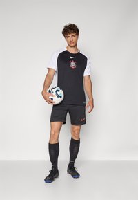 Tricou sportiv alb-negru cu un logo grafic, asortat cu pantaloni scurți negri ce prezintă un logo roșu Nike. Jucătorul ține o minge de fotbal cu un model.