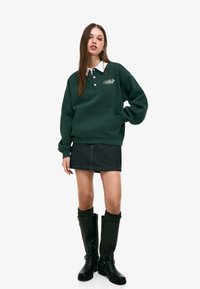 Temno zelena sweatshirt s belim ovratnikom, sprednjim žepom in detajlom logotipa. V kombinaciji z denarnico in črnimi škornji do kolen.