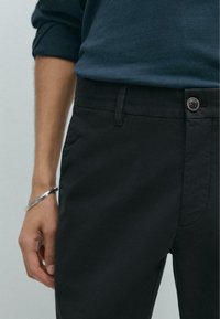 Schwarze Baumwollhosen mit glatter Textur, ausgestattet mit einem Knopfverschluss, Gürtelschlaufen und schlanker Passform, kombiniert mit einem dunkelblauen Pullover.