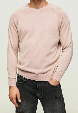 Mężczyzna w jasnoróżowym swetrze z długim rękawem i czarnych podartych dżinsach, z jedną ręką w kieszeni, pokazany od szyi do bioder.