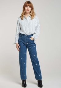 Chemise à rayures blanche et bleu clair, portée avec un jean en denim bleu arborant des broderies florales et une coupe droite. Bottines noires.