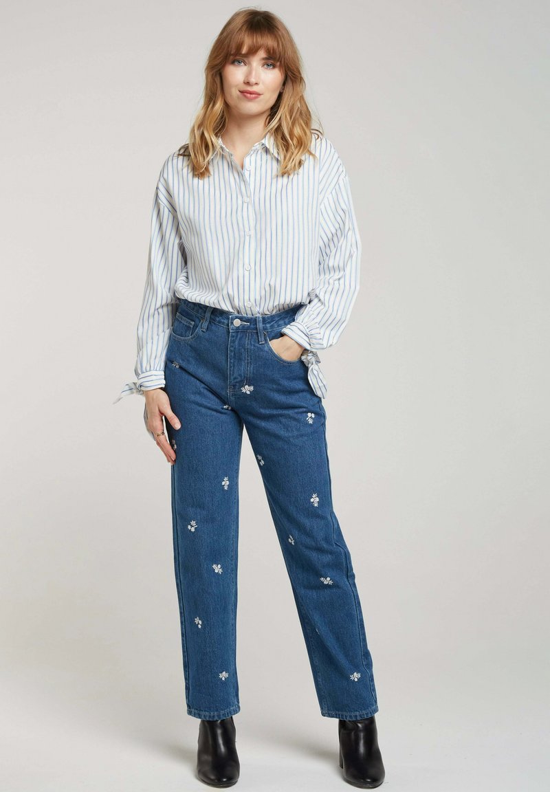 Chemise à rayures blanche et bleu clair, portée avec un jean en denim bleu arborant des broderies florales et une coupe droite. Bottines noires.