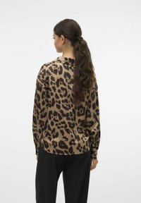 Blusa con stampa leopardata in beige e marrone, maniche lunghe, texture liscia, orlo arrotondato, indossata con pantaloni neri, che mette in mostra una vestibilità rilassata.